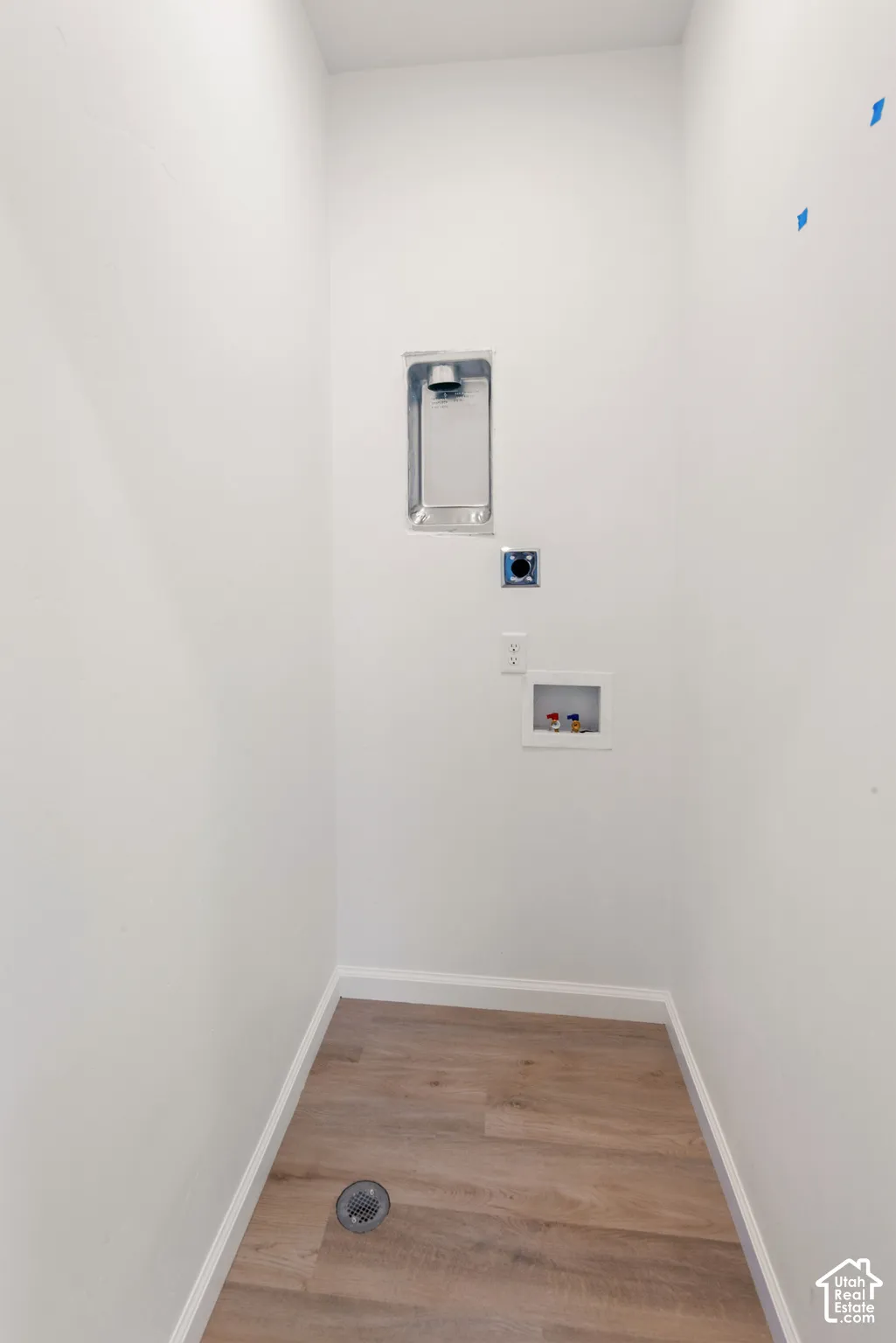 Utility space featuring wood-finish flooring, a recessed wall-mounted dryer vent, an electrical outlet, and a recessed plumbing connection box