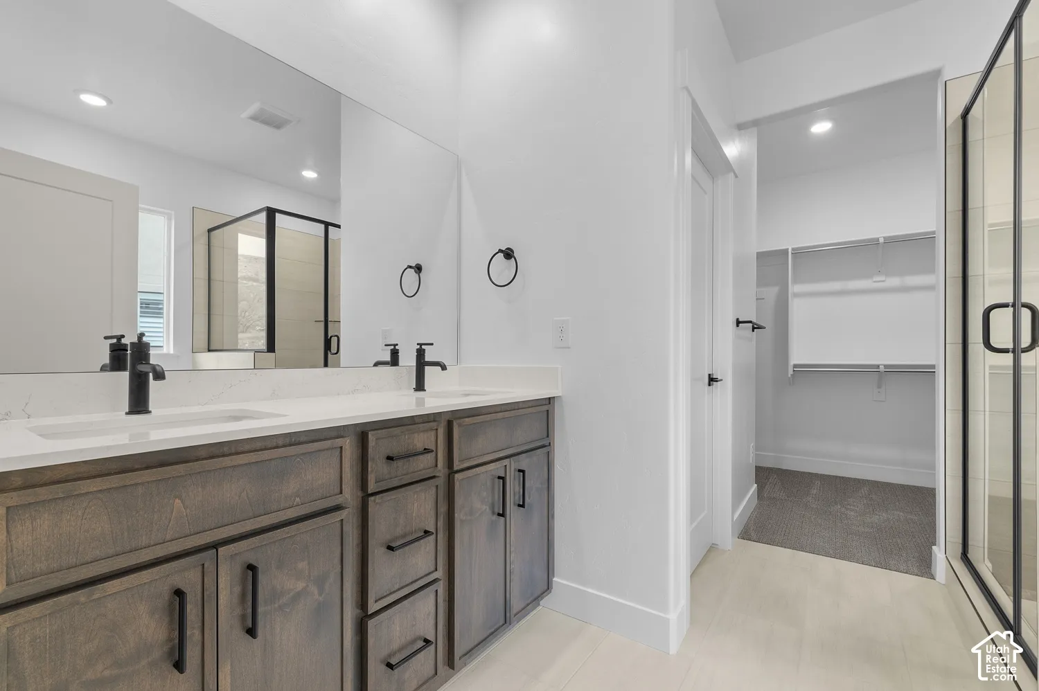 Dual-sink vanity with stone-style countertops, dark wood-finish cabinetry, matte black hardware, expansive wall-to-wall mirror, and a walk-in glass shower enclosure