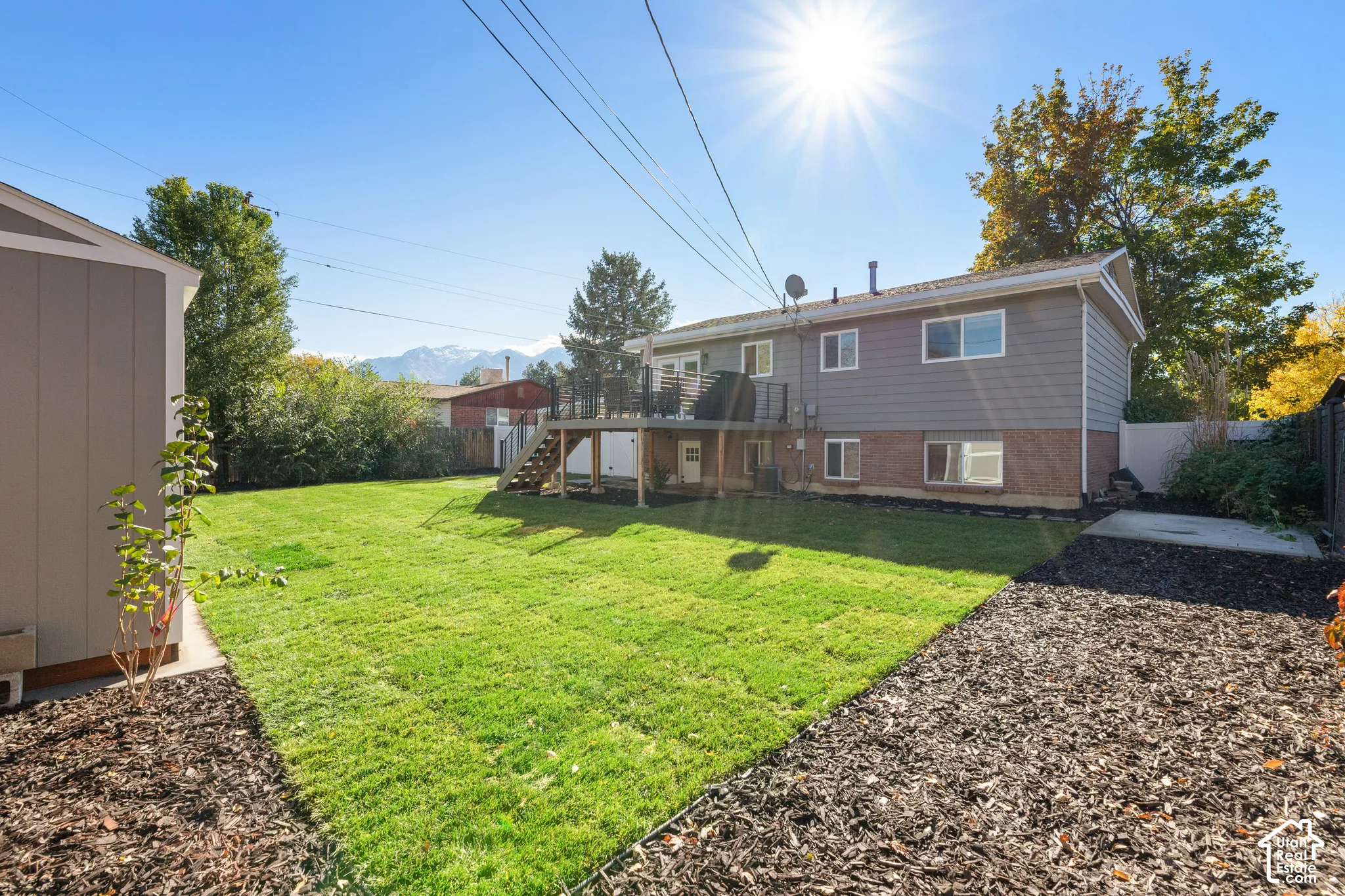 Expansive backyard featuring a manicured lawn, mulched garden beds, a raised wood deck with metal railing, and a detached storage structure, set against a backdrop of mountains