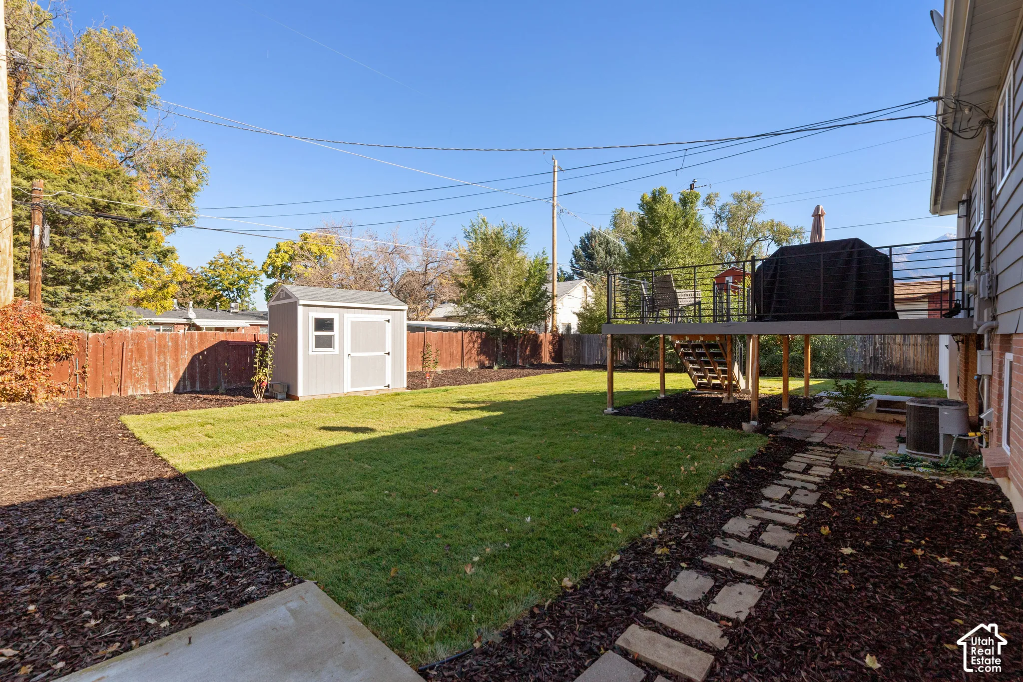 Fenced backyard featuring a raised composite deck with metal railing, an elevated storage shed, stone paver walkway, and mulched landscaping