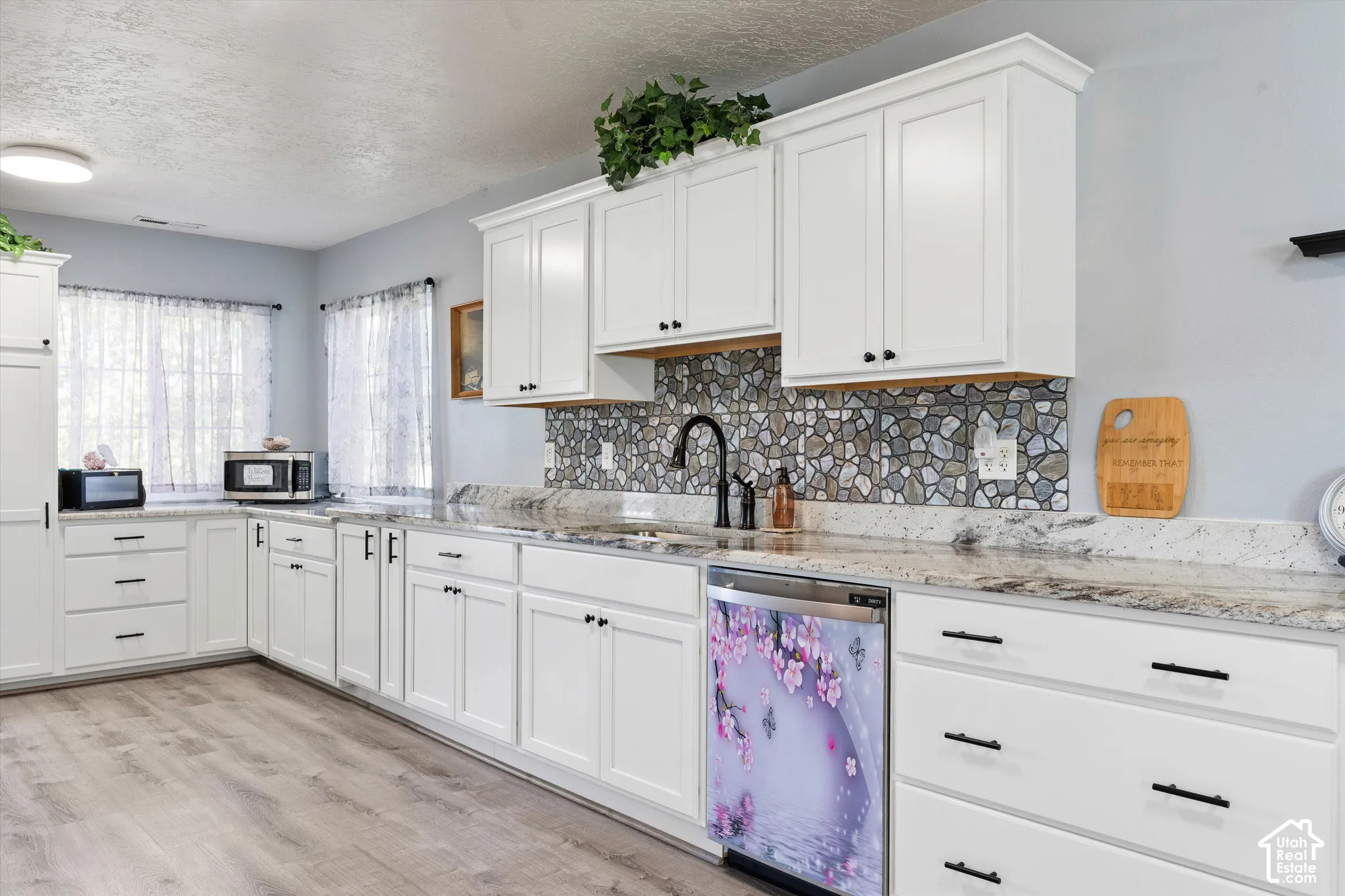 Kitchen featuring white shaker-style cabinetry, stone-finish countertops, decorative pebble-style tile backsplash, black hardware, and wood-finish flooring