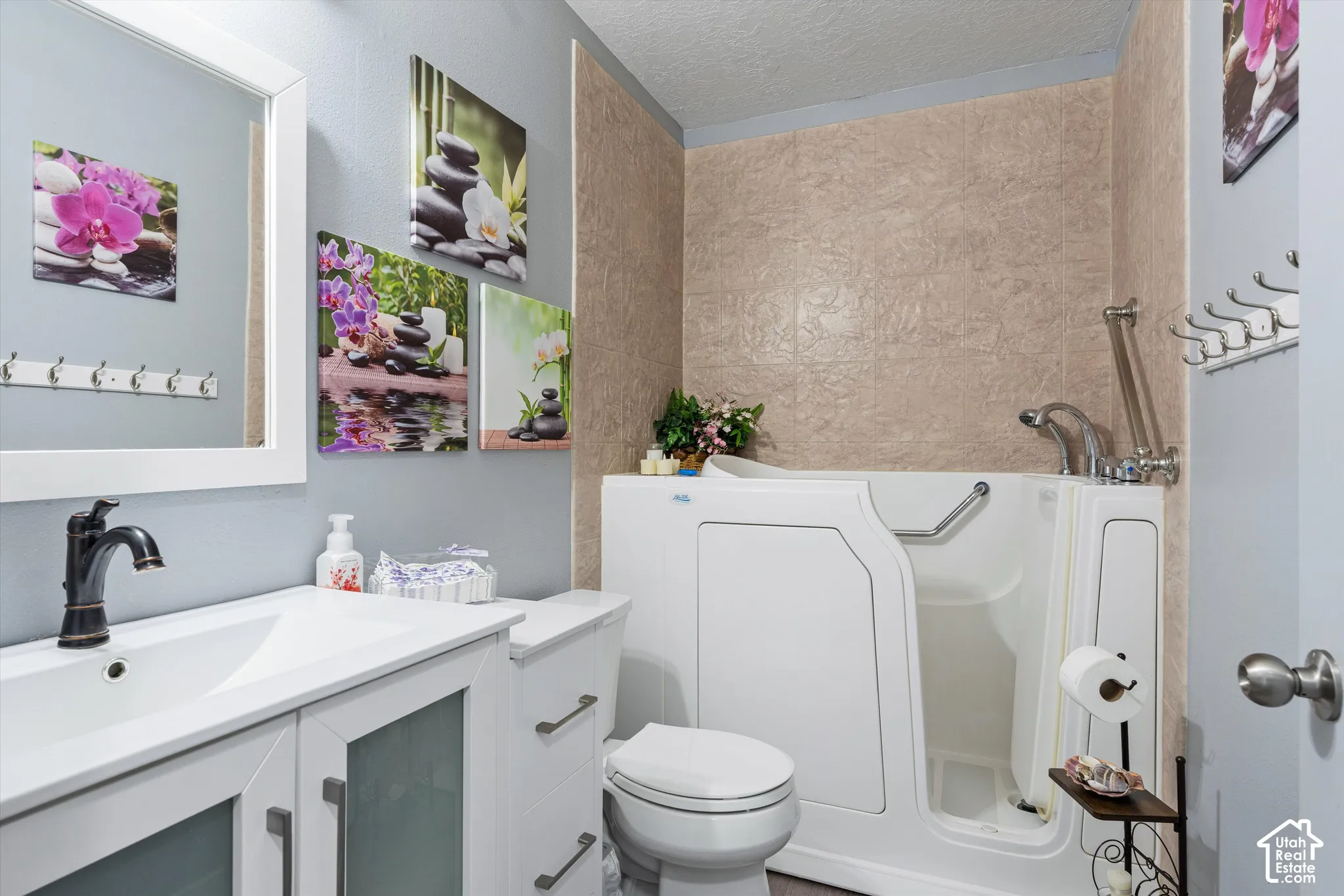 Bathroom featuring a white vanity with frosted glass cabinet inserts, a rectangular basin with a dark metal faucet, a step-in soaking tub with a door, and textured surround wall cladding