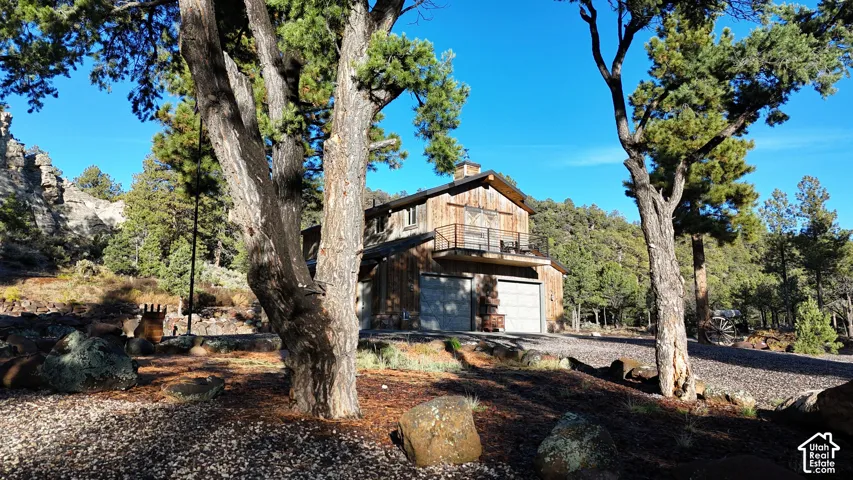 Two-story wood-sided exterior featuring an upper-level metal-railing balcony and a multi-car garage with overhead doors, situated on a rock-landscaped lot with mature trees and native terrain