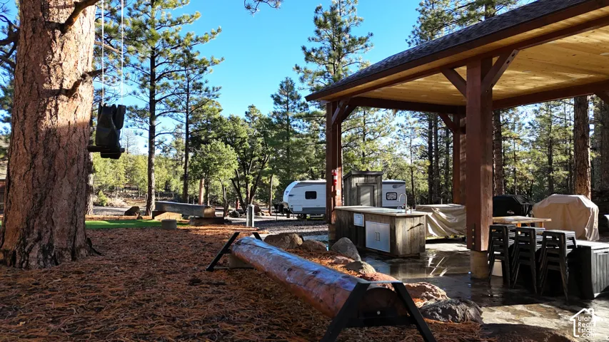 Covered timber-frame outdoor structure featuring a stone patio, built-in kitchen area, log bench, and mature pine trees
