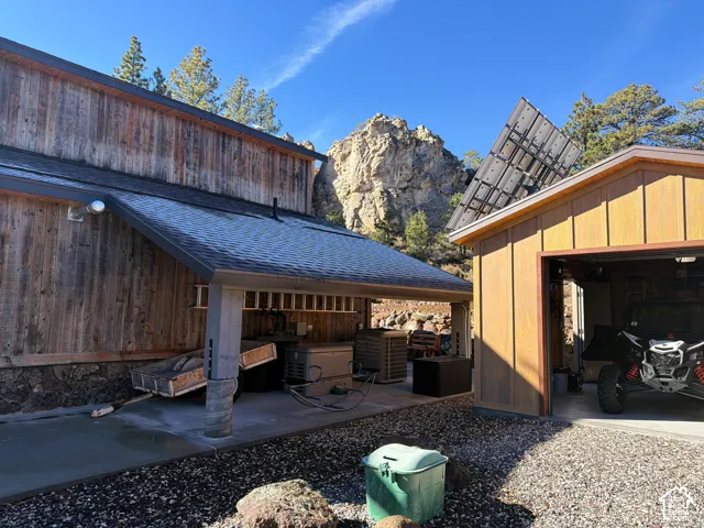 Vertical wood siding exterior with a covered concrete patio, detached garage, and solar panel array set against a prominent rock formation