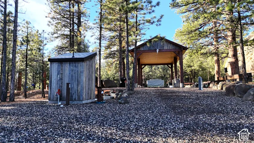 Outdoor area featuring a wood-sided utility structure, a timber-framed pavilion with a vaulted ceiling, gravel grounds, and mature pine trees