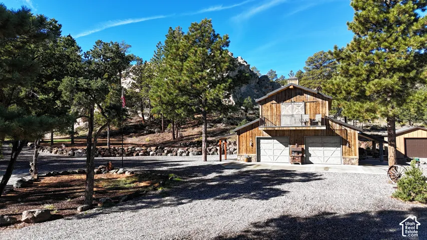 Rustic wood-sided structure featuring an upper-level balcony, two ground-level garage bays, stone foundation detailing, and an attached side carport