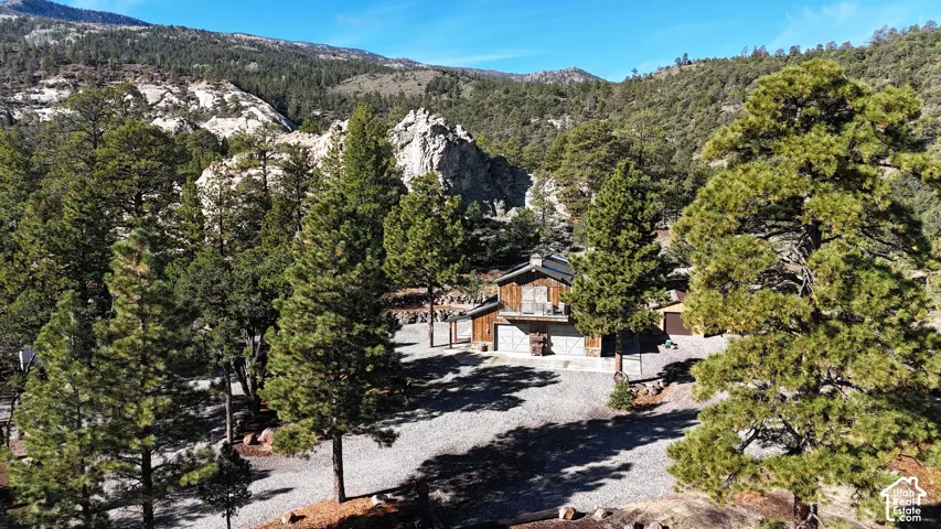 Wood-sided structure featuring an upper-level balcony, a stone chimney, a detached garage with dual carriage-style doors, and extensive evergreen tree coverage set against a mountainous landscape