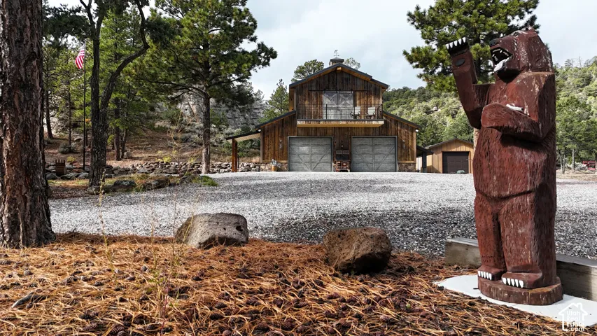 Rustic wood-paneled exterior featuring two metal garage doors, a second-story balcony with metal railings, a stone foundation, and a gravel driveway