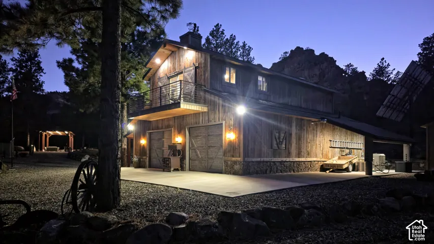 Rustic wood-clad exterior with stone foundation, second-story balcony featuring metal railing, dual garage bays, integrated exterior lighting, and solar array backdrop