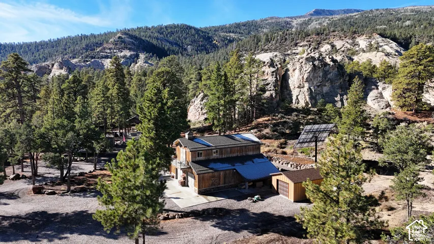 Rustic two-story wood-sided structure featuring a metal roof, attached garage, separate outbuilding, and elevated solar array, set against a mountainous, forested landscape