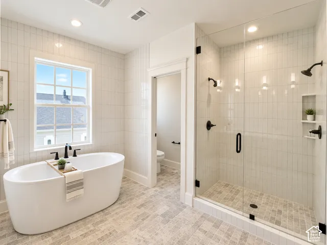 Freestanding soaking tub with a wall-mounted faucet, walk-in glass-enclosed shower with dual shower heads, integrated recessed shelving, and porcelain tile wall detailing