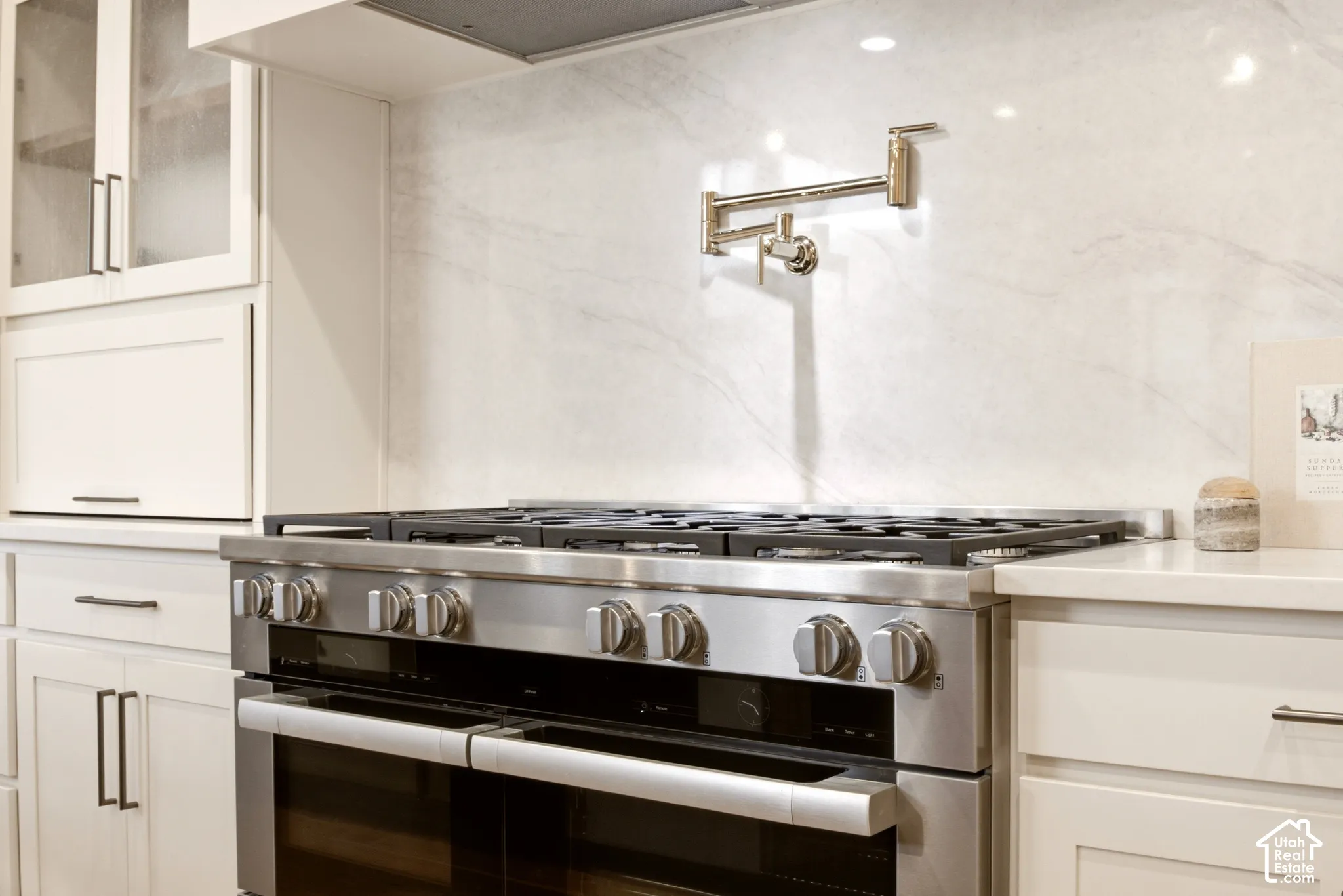 Miele 48" dual fuel double-oven range, Ivory Taj quartz backsplash, wall-mounted Kohler French Gold pot filler, custom appliance garage cabinetry with "Kokomo Seedy" glass-panel upper doors, and Ivory Taj Quartz countertop