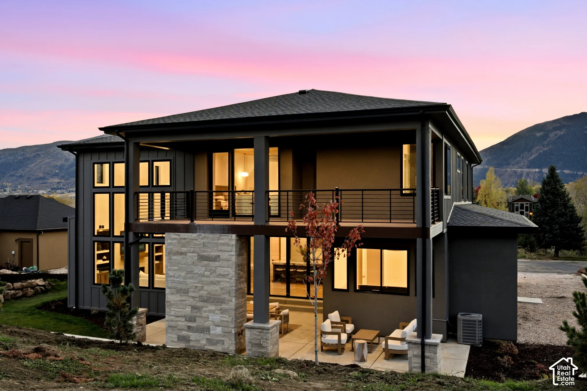 Multi-level exterior with expansive windows, natural limestone fireplace, covered patio, private primary balcony with horizontal rail system, and mountain backdrop