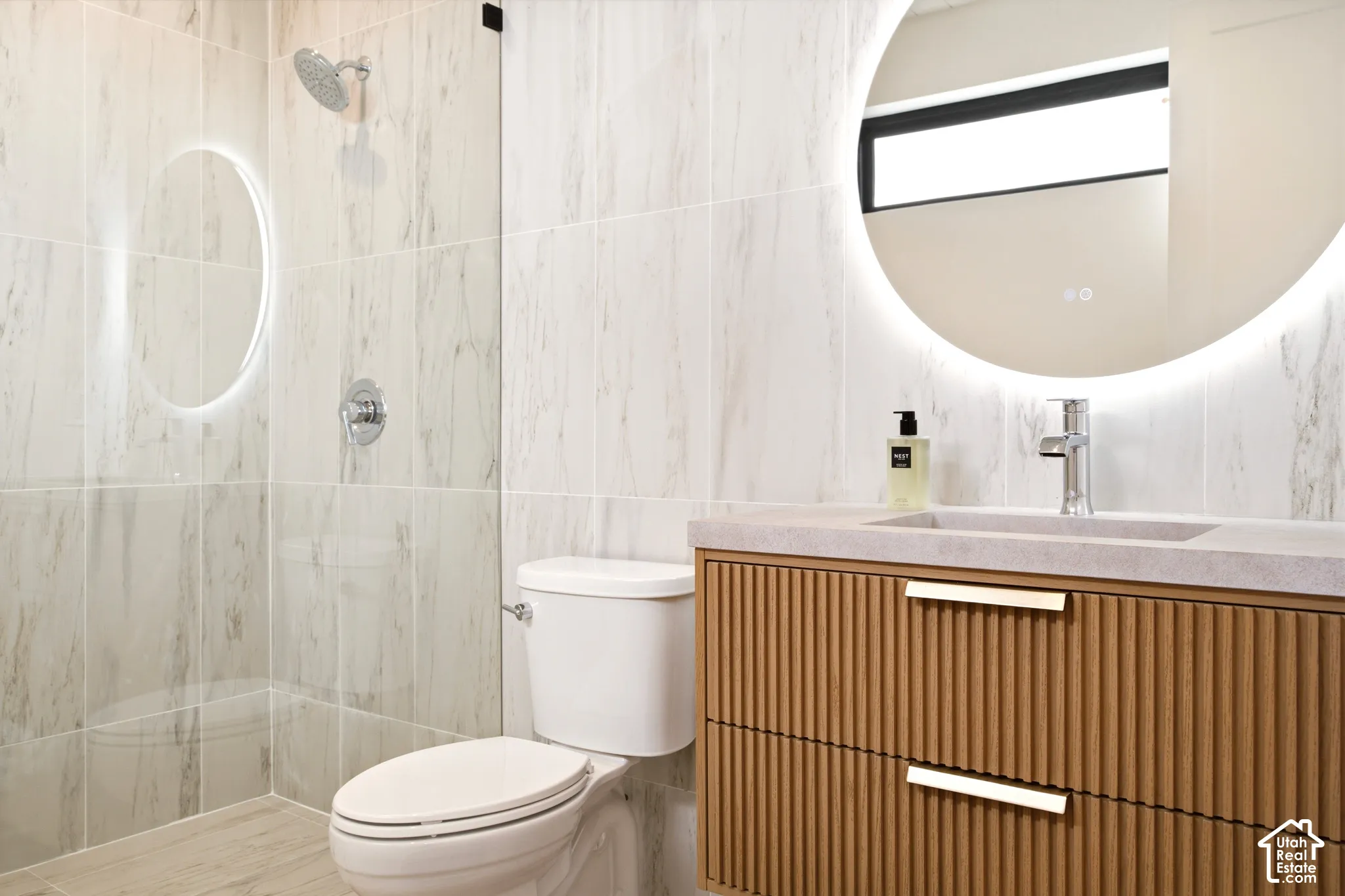 En-Suite Bathroom featuring floor-to-ceiling Calcutta large-format tile, a fluted floating wood vanity with a solid-surface countertop, an integrated sink with a chrome faucet, a frameless glass shower with a rainfall showerhead, and a backlit circular mi