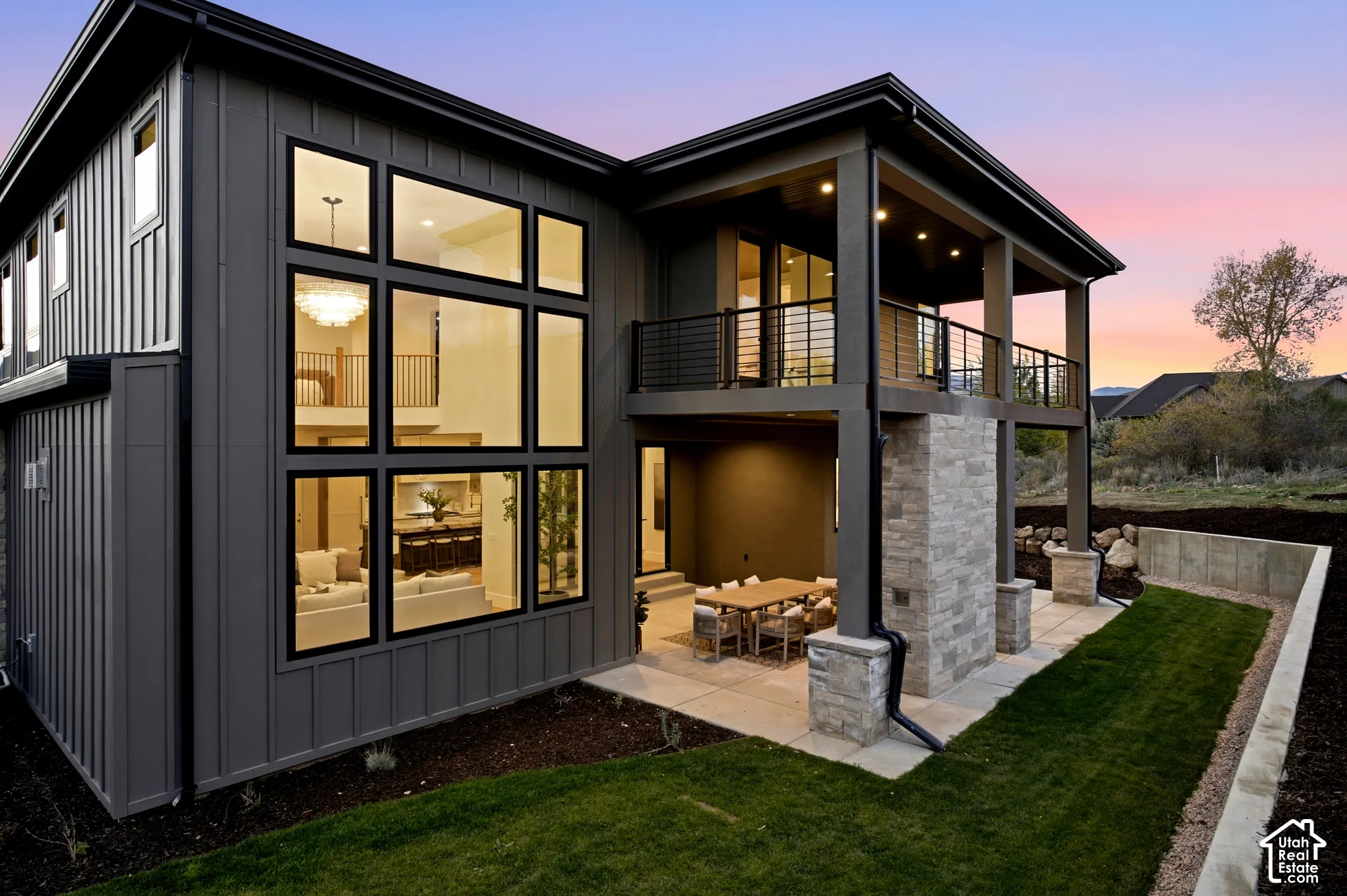 Two-story exterior featuring board-and-batten siding, large-format glass windows, dry-stacked natural limestone fireplace, a concrete patio, and a covered balcony with metal railings