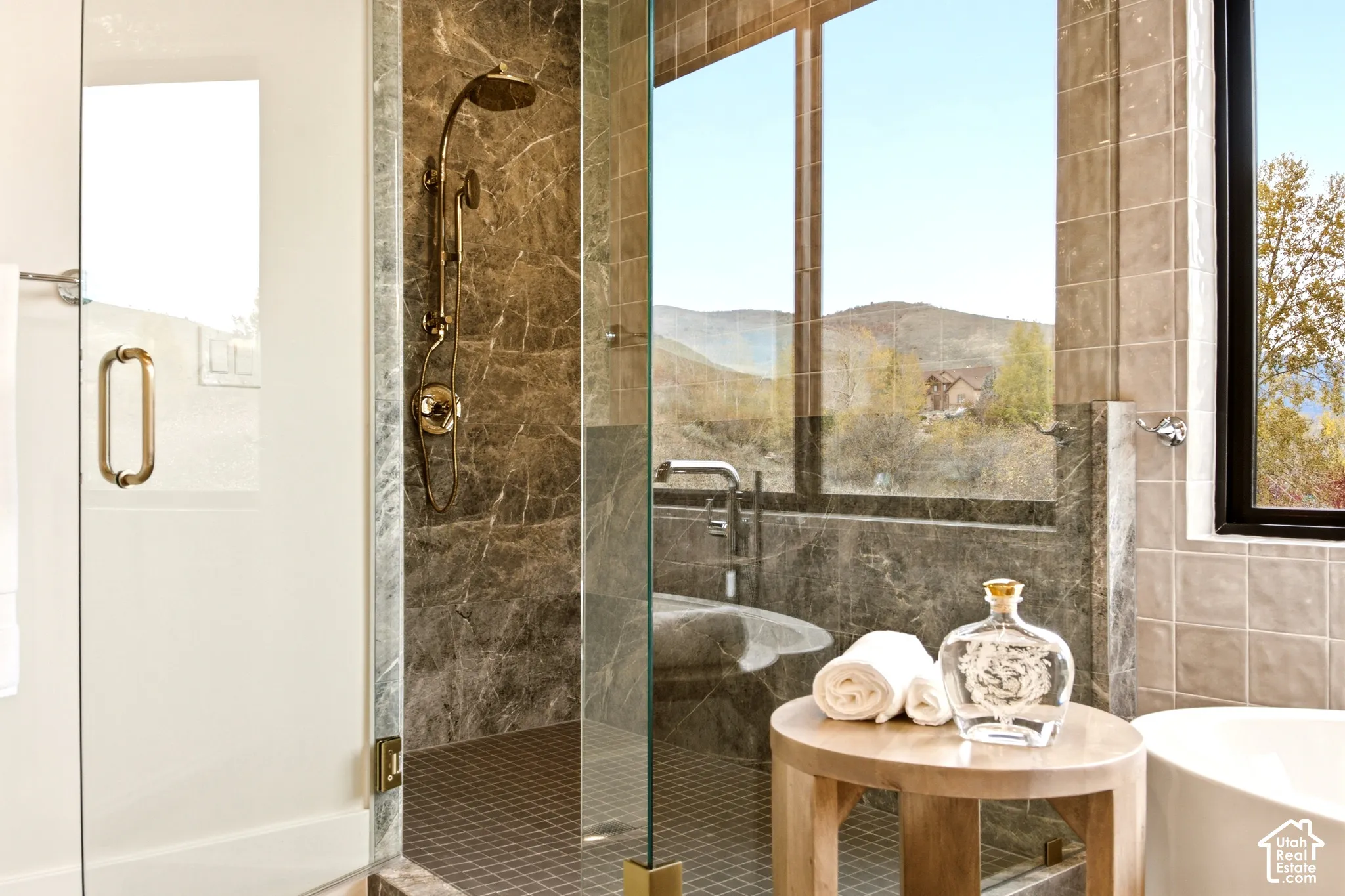 Walk-in jewel-box shower featuring marble look Edward Martin wall tile, Kohler French Gold rainfall shower, accompanied by a freestanding soaking tub set against expansive windows