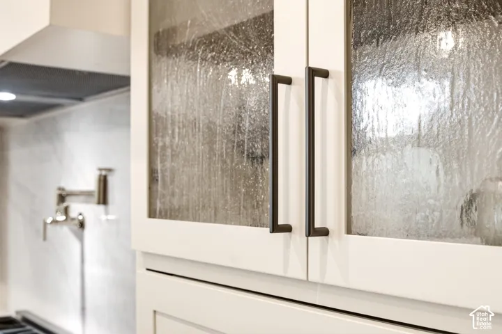 Creamy white cabinetry featuring"Kokomo Seedy" glass inserts, ash-grey hardware, and a Kohler French gold,wall-mounted pot filler over Ivory Taj- Quartz backsplash