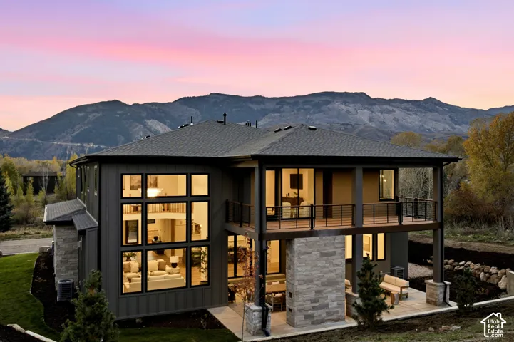 Multi-level exterior with expansive windows, natural limestone fireplace, covered patio, private primary balcony with horizontal rail system, and mountain backdrop