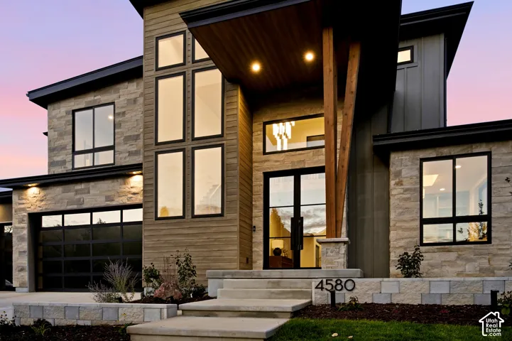 Modern exterior featuring natural limestone siding, horizontal wood-finish siding, full view tinted glass garage doors, multi-pane glass entryway, and a statement timber support