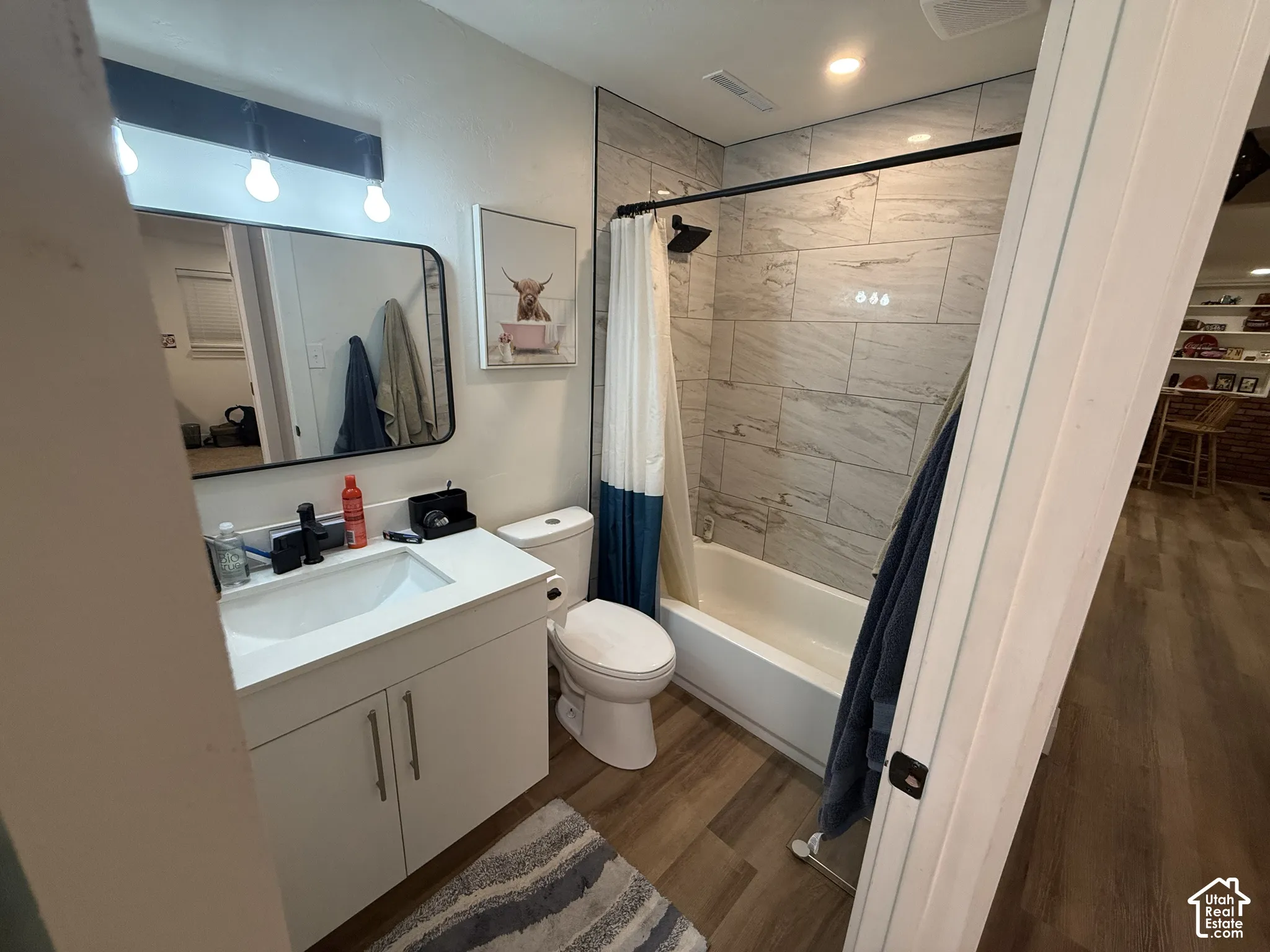 Bathroom featuring a white vanity with an integrated basin, a black wall-mounted vanity light, wood-finish flooring, and a shower-tub combination with horizontal wall tiling