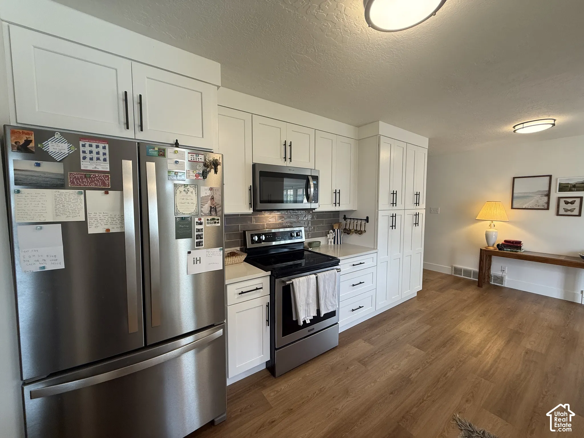 Kitchen featuring white shaker-style cabinetry, stainless steel appliances, a grey tiled backsplash, light-toned countertops, and wood-finish flooring
