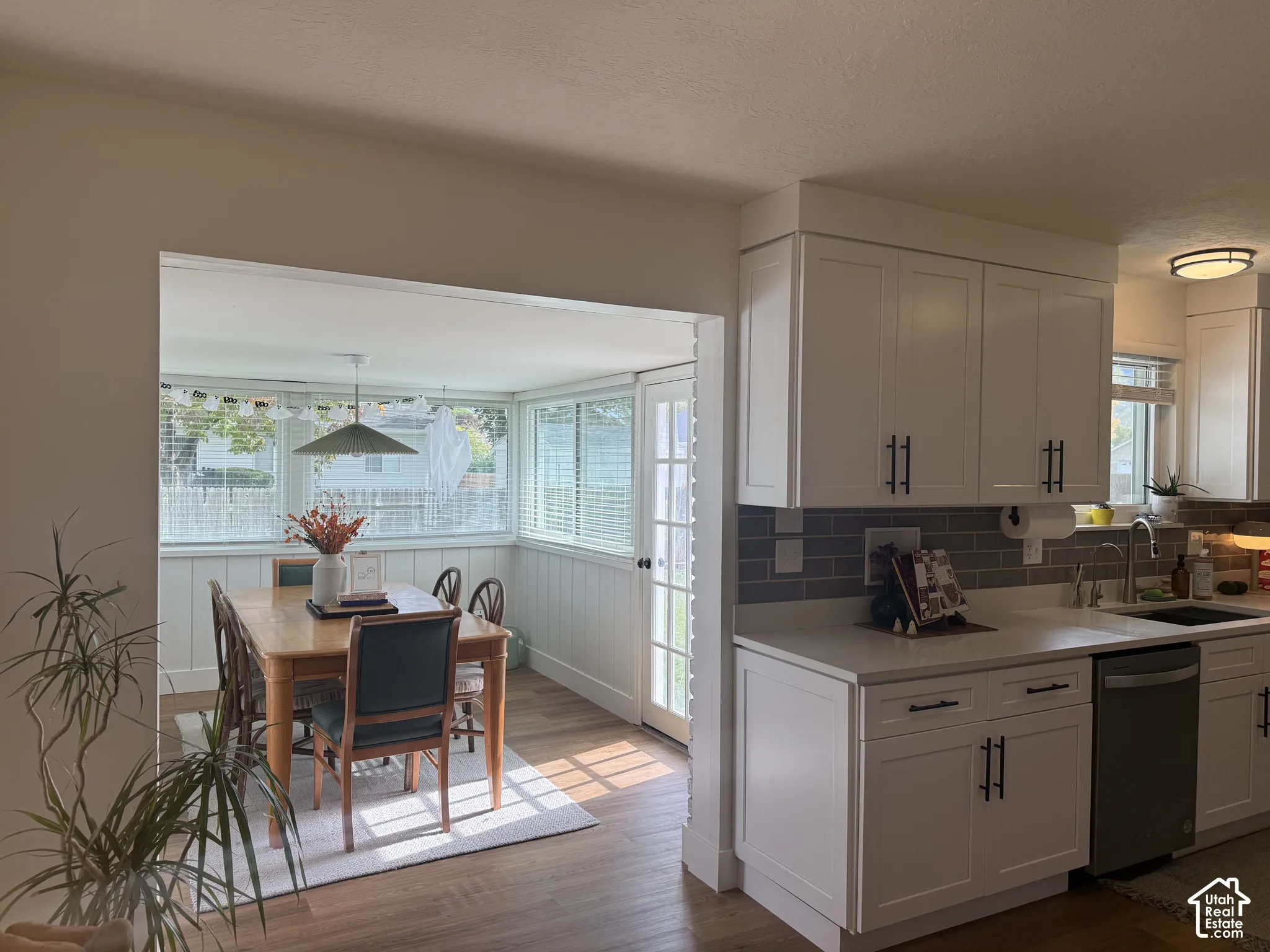 Kitchen featuring white Shaker-style cabinetry, dark tile backsplash, light-colored countertops, and stainless steel dishwasher, complemented by wood-finish flooring