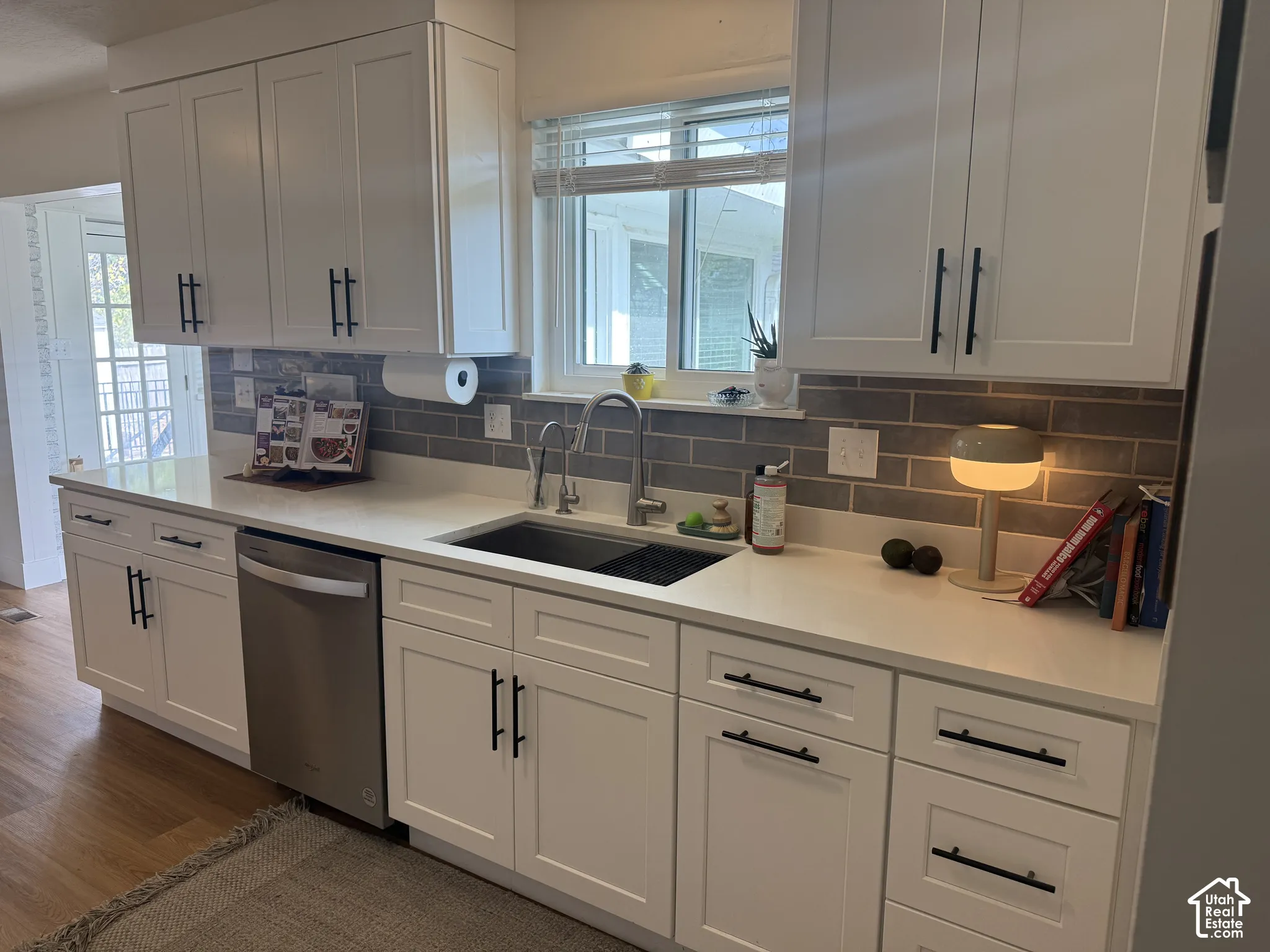 White Shaker-style cabinetry featuring matte black hardware, stone-composite countertops, horizontal grey subway tile backsplash, stainless steel undermount sink, and stainless steel dishwasher