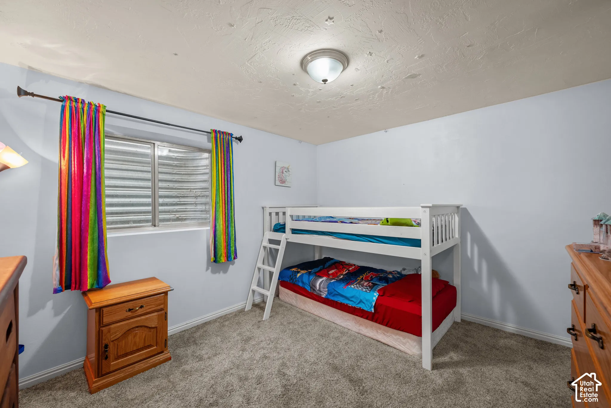 Bedroom featuring neutral-toned carpeting, textured ceiling, flush-mount lighting, and an egress window with a metal window well