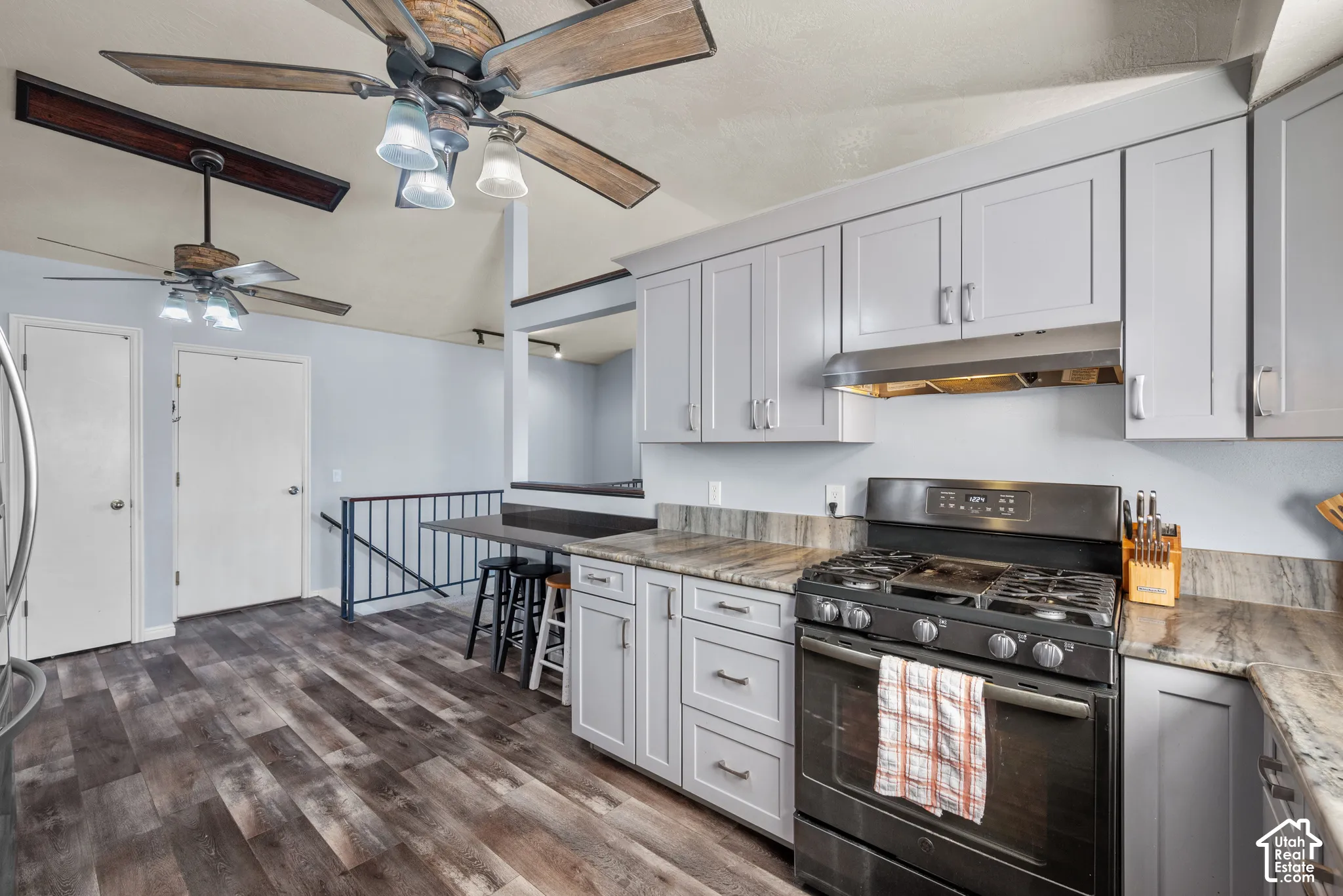 Kitchen featuring light grey Shaker-style cabinetry, stone-patterned countertops, a stainless steel gas range with overhead exhaust, and wood-finish flooring, complemented by vaulted ceilings with integrated ceiling fans