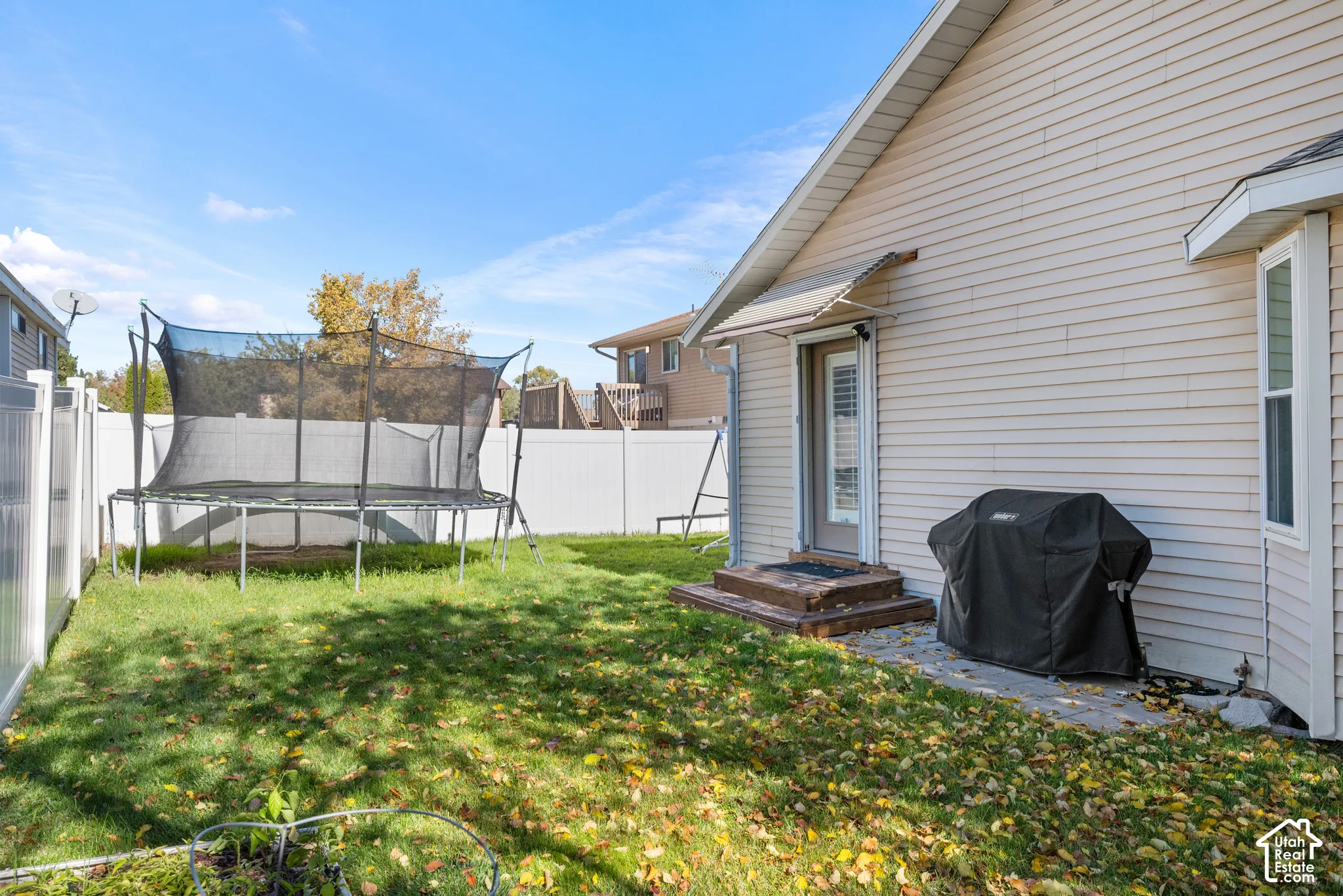 Rear exterior featuring light-colored horizontal siding, white vinyl privacy fencing, a stone-paved patio, and a metal door awning
