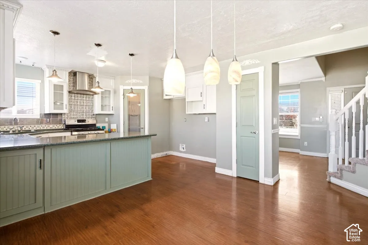 Kitchen featuring wood-finish flooring, a central island with beadboard cabinetry, stainless steel range hood, glass-front upper cabinetry, and trio of pendant light fixtures