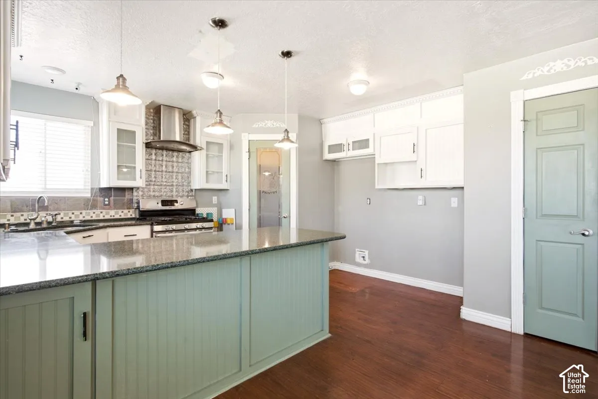 Kitchen featuring a peninsula with a stone-finish countertop, white cabinetry with glass-front uppers, stainless steel range hood, pendant lighting, and wood-finish flooring