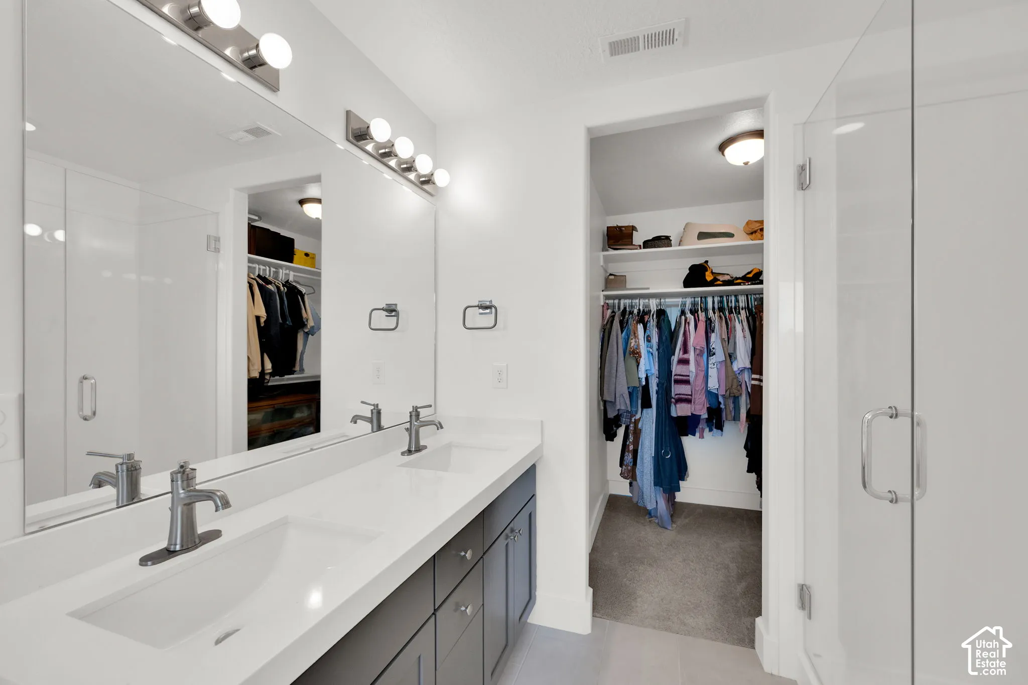 Dual-sink vanity with white composite countertops, large wall-to-wall mirror, glass-enclosed shower, and walk-in closet with built-in shelving