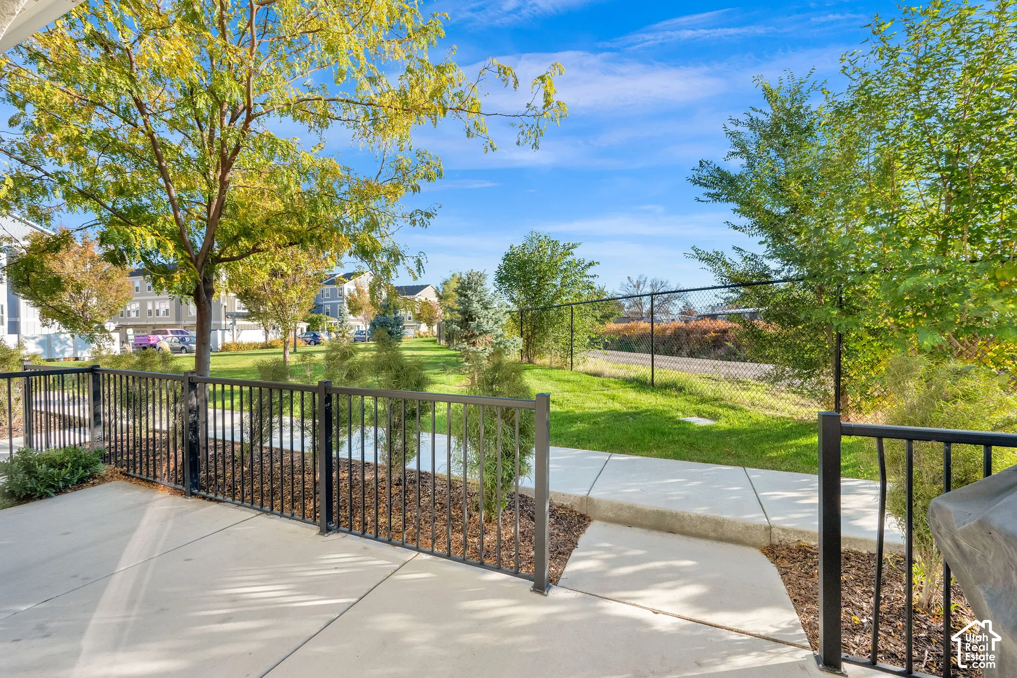 Concrete patio featuring a black metal railing, paved walkway, manicured lawn, and mature trees enclosed by perimeter fencing
