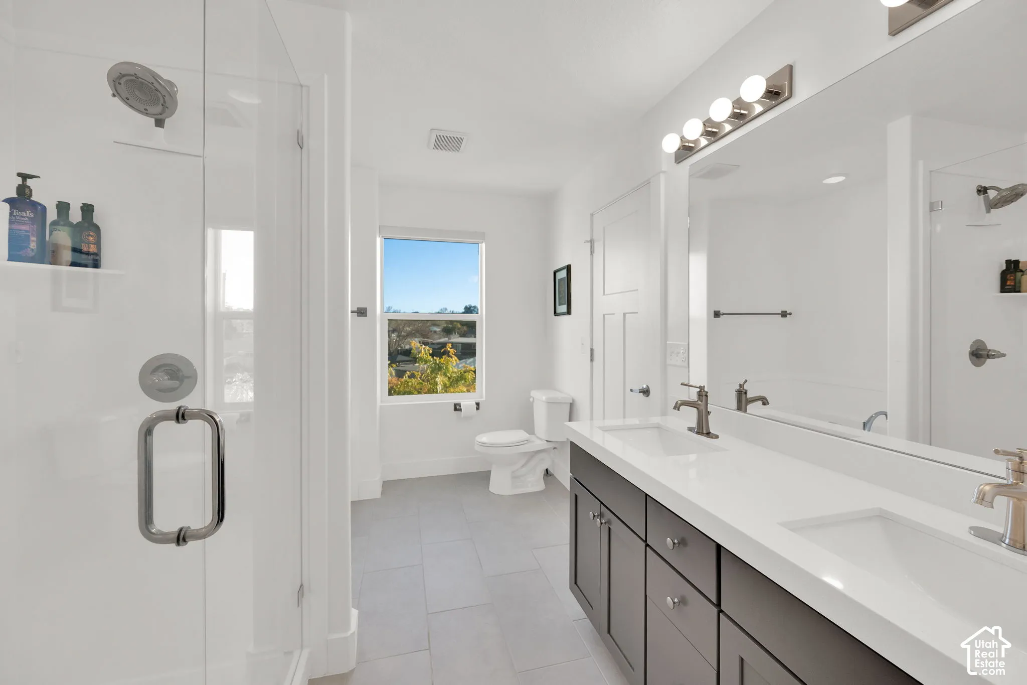 Bathroom featuring a dual-sink vanity with white countertops, gray cabinetry, a frameless glass-enclosed shower, and large-format tile flooring