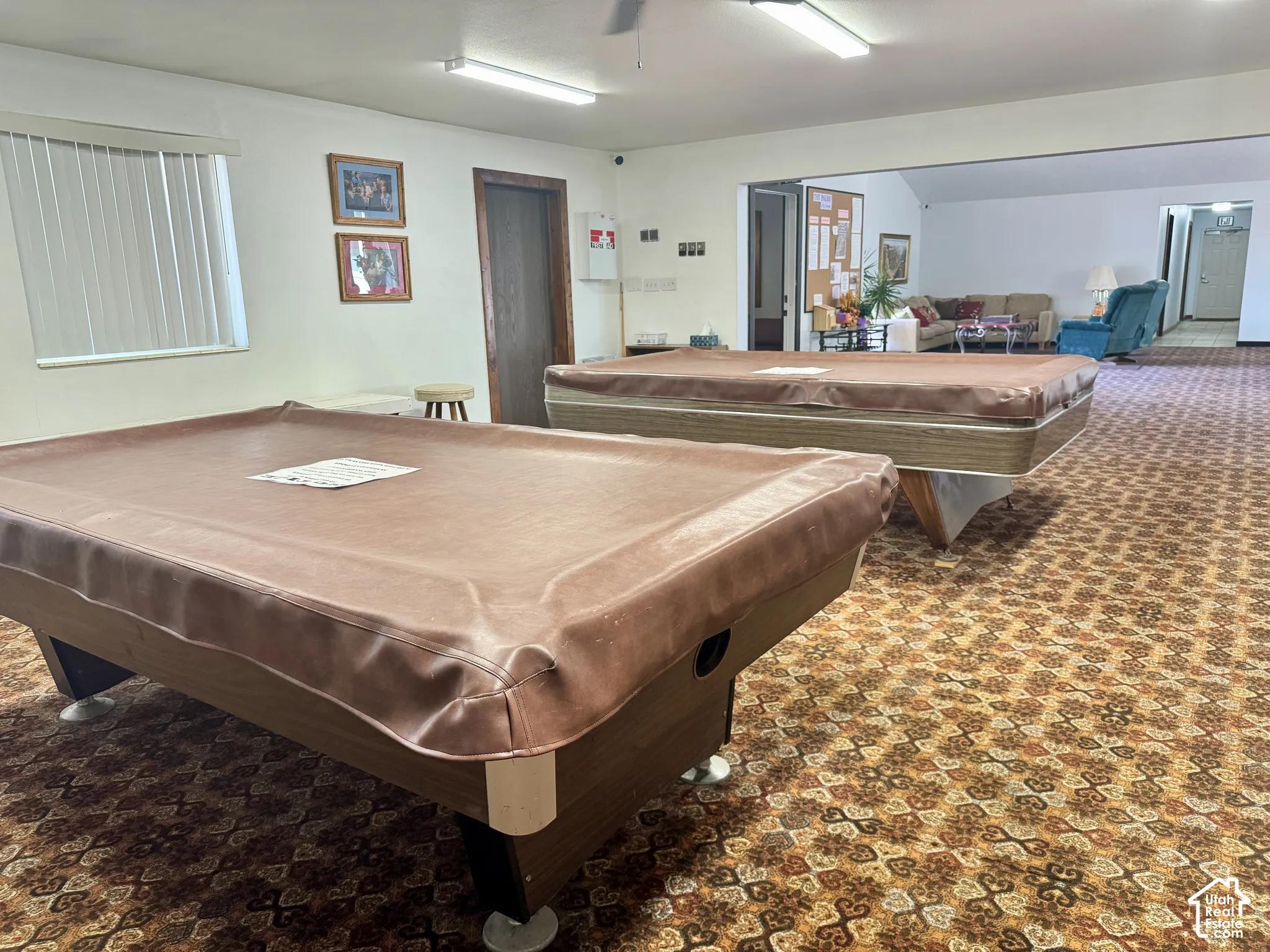 Expansive interior space featuring patterned wall-to-wall carpeting, linear fluorescent ceiling lighting, and horizontal slatted window blinds