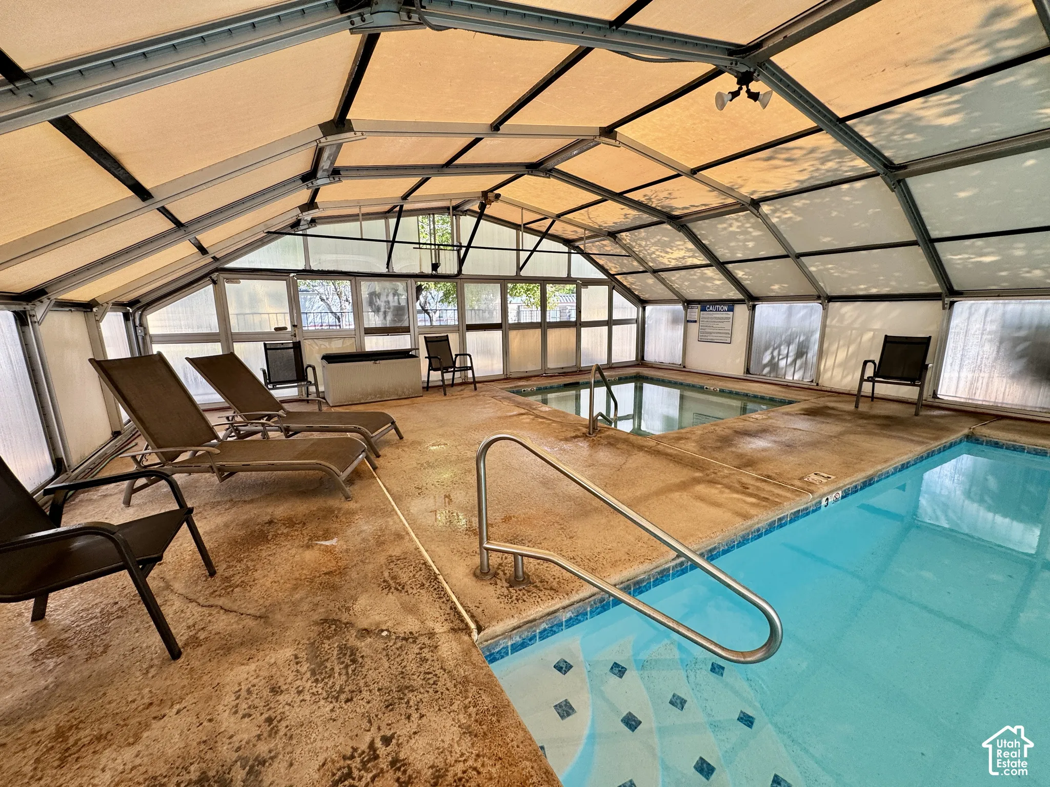 Enclosed swimming pool area featuring a metal-framed structure with a translucent roof, a dedicated hot tub, concrete decking, and stainless steel handrails
