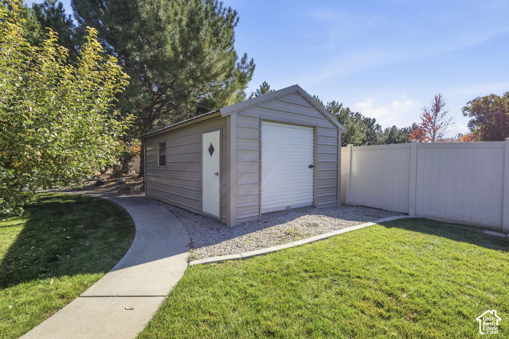 Detached metal storage structure featuring a roll-up garage door, side entry door with a diamond-shaped window, and concrete walkway set against a vinyl privacy fence