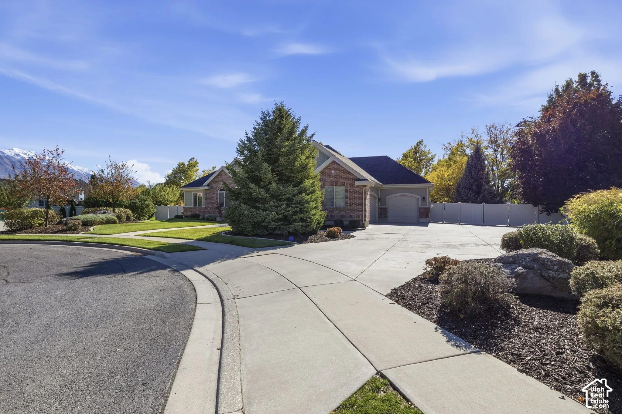 Brick exterior with multi-gabled roofline, concrete driveway, attached garage, and manicured landscaping featuring mature trees and mountain backdrop