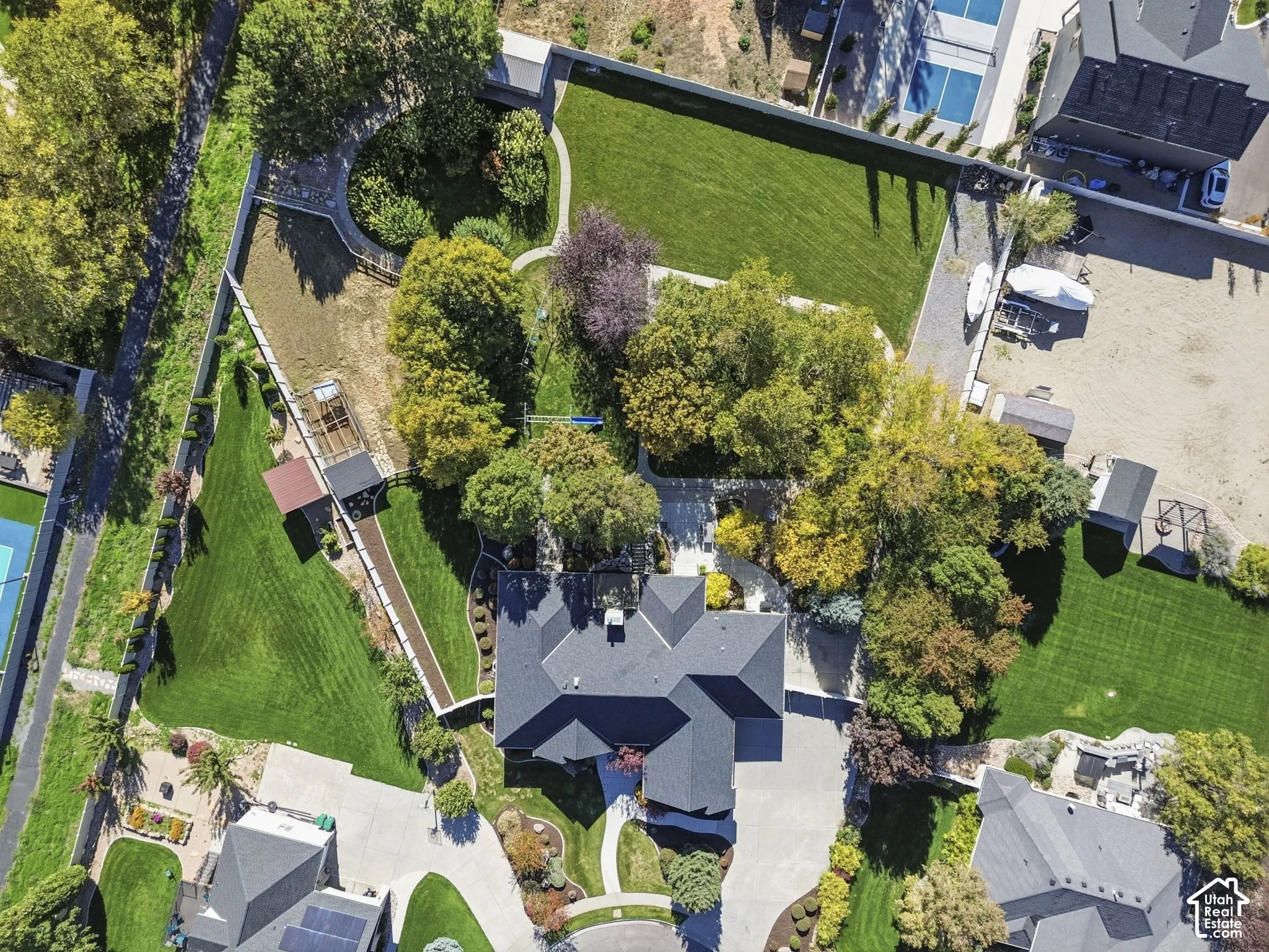 Aerial view featuring a multi-gabled roofline, expansive manicured lawn, mature trees, concrete driveway, and fenced perimeter