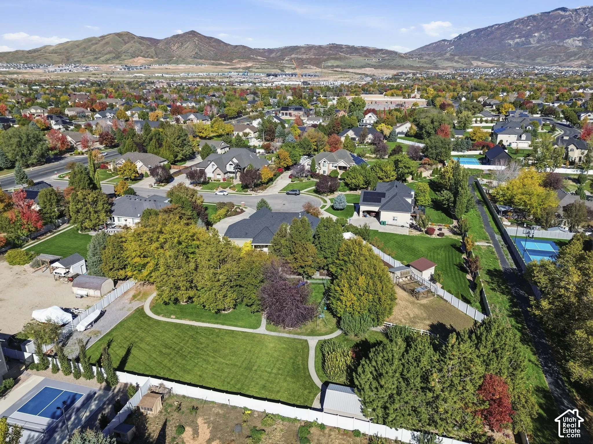 Aerial perspective featuring expansive manicured lawns, mature shade trees, white vinyl perimeter fencing, and mountain scenery