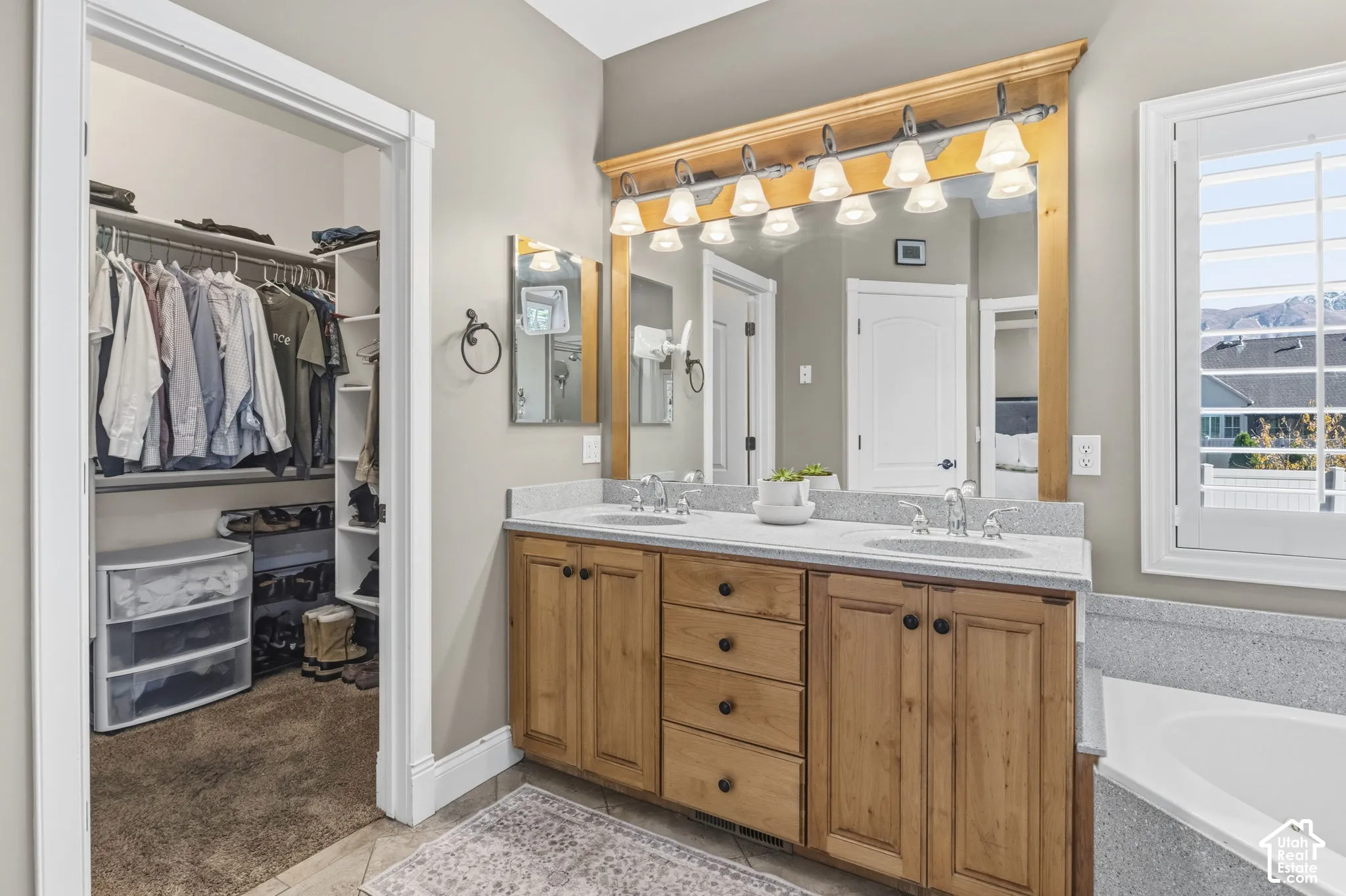 Dual-sink vanity with wood cabinetry, light-toned stone-finish countertop, full-width mirror with wood frame, multi-light fixture, and walk-in closet with built-in shelving
