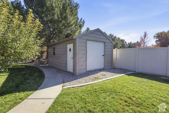 Detached metal storage structure featuring a roll-up garage door, side entry door with a diamond-shaped window, and concrete walkway set against a vinyl privacy fence