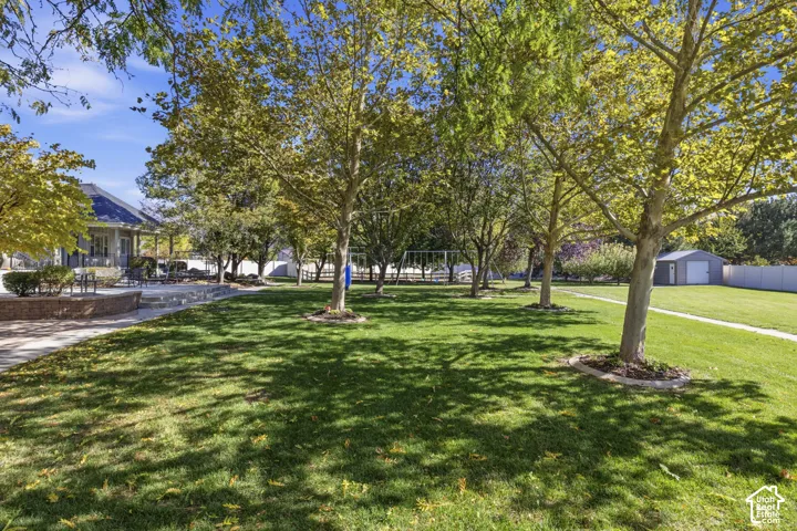 Expansive grounds featuring a manicured lawn, mature deciduous trees, a brick-retaining wall patio, and a detached storage structure enclosed by perimeter fencing