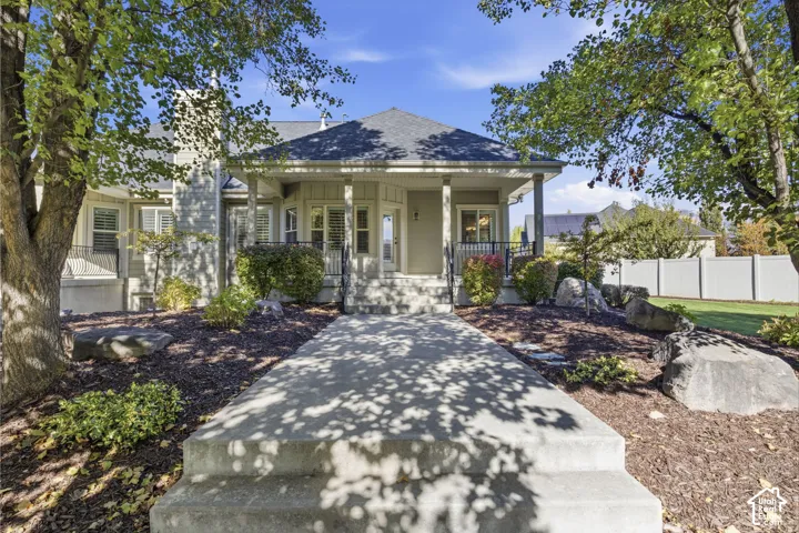 Front exterior featuring a covered porch with decorative metal railing, concrete steps, landscaped grounds with mature trees and large boulders, and a neutral siding finish