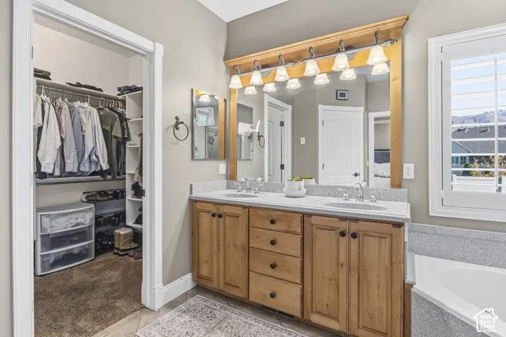 Dual-sink vanity with wood cabinetry, light-toned stone-finish countertop, full-width mirror with wood frame, multi-light fixture, and walk-in closet with built-in shelving