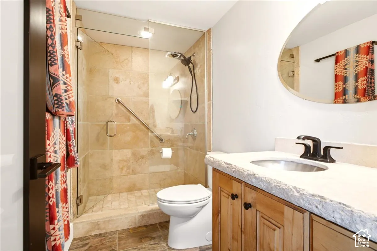 Bathroom featuring a natural stone countertop with a chiseled edge, a wood-finish vanity cabinet, a walk-in shower with stone tile surround and a glass door, and an oval wall-mounted mirror