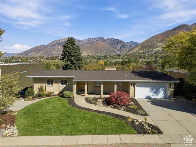 Single-story exterior featuring a brick facade, covered entryway, attached two-car garage, manicured lawn with landscape beds, and a prominent mountain backdrop