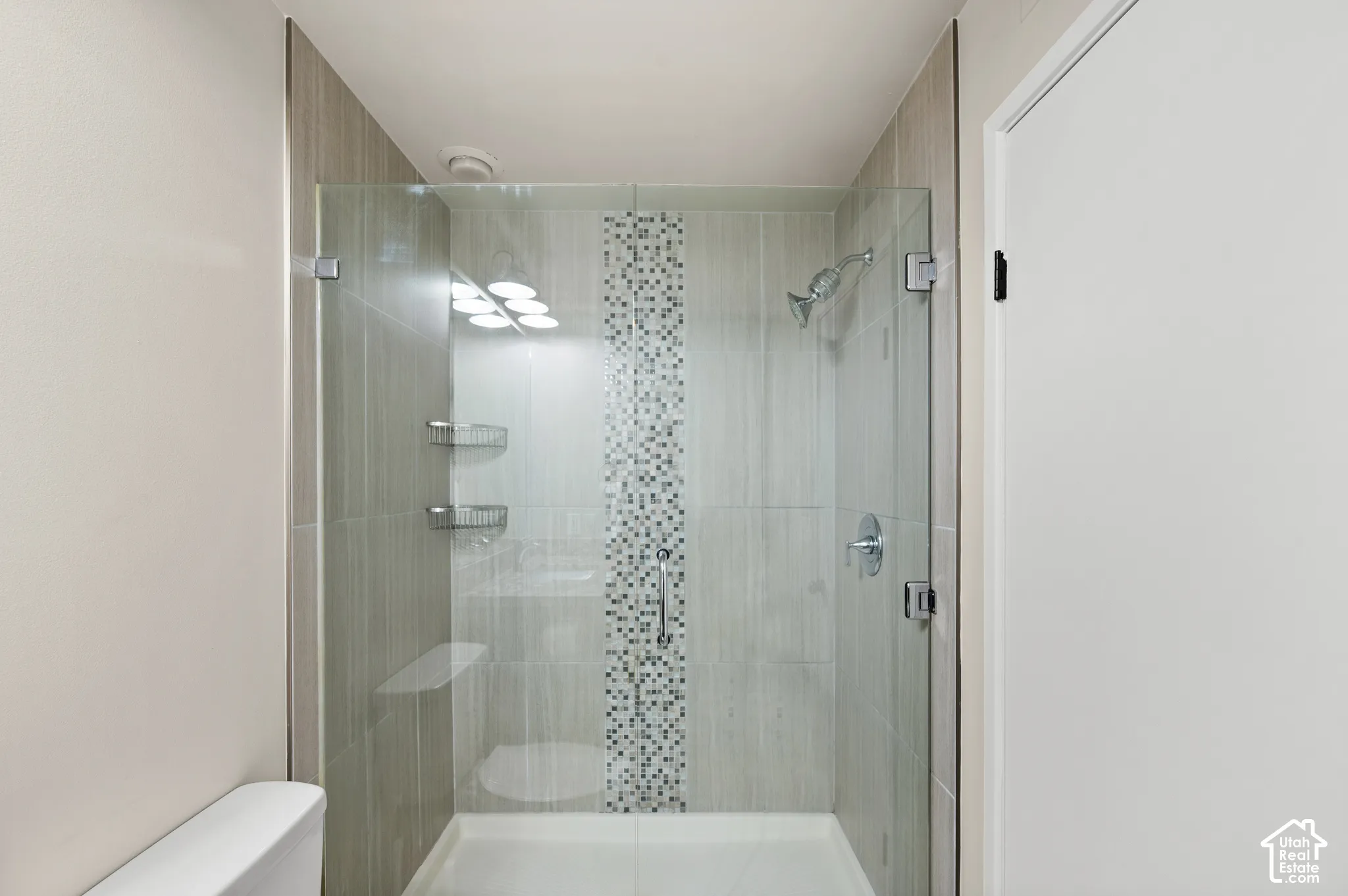 Walk-in shower featuring glass paneling, floor-to-ceiling neutral tile, a decorative vertical mosaic accent strip, wall-mounted metal shelving, and a polished metal shower head